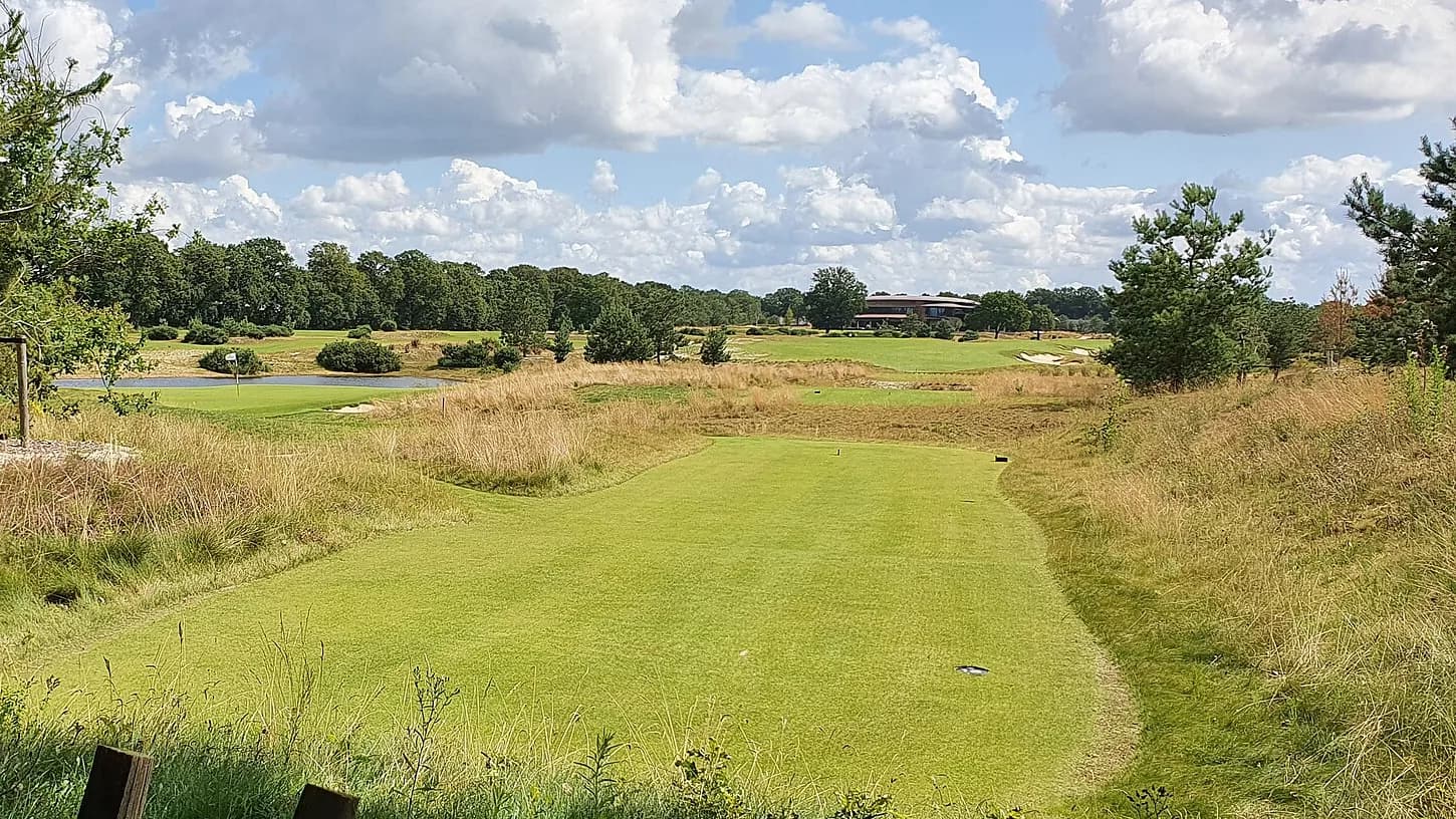 Een tee tussen hoog gras op Bernardus Golf, met in de verte een vijver, de green en het clubhuis