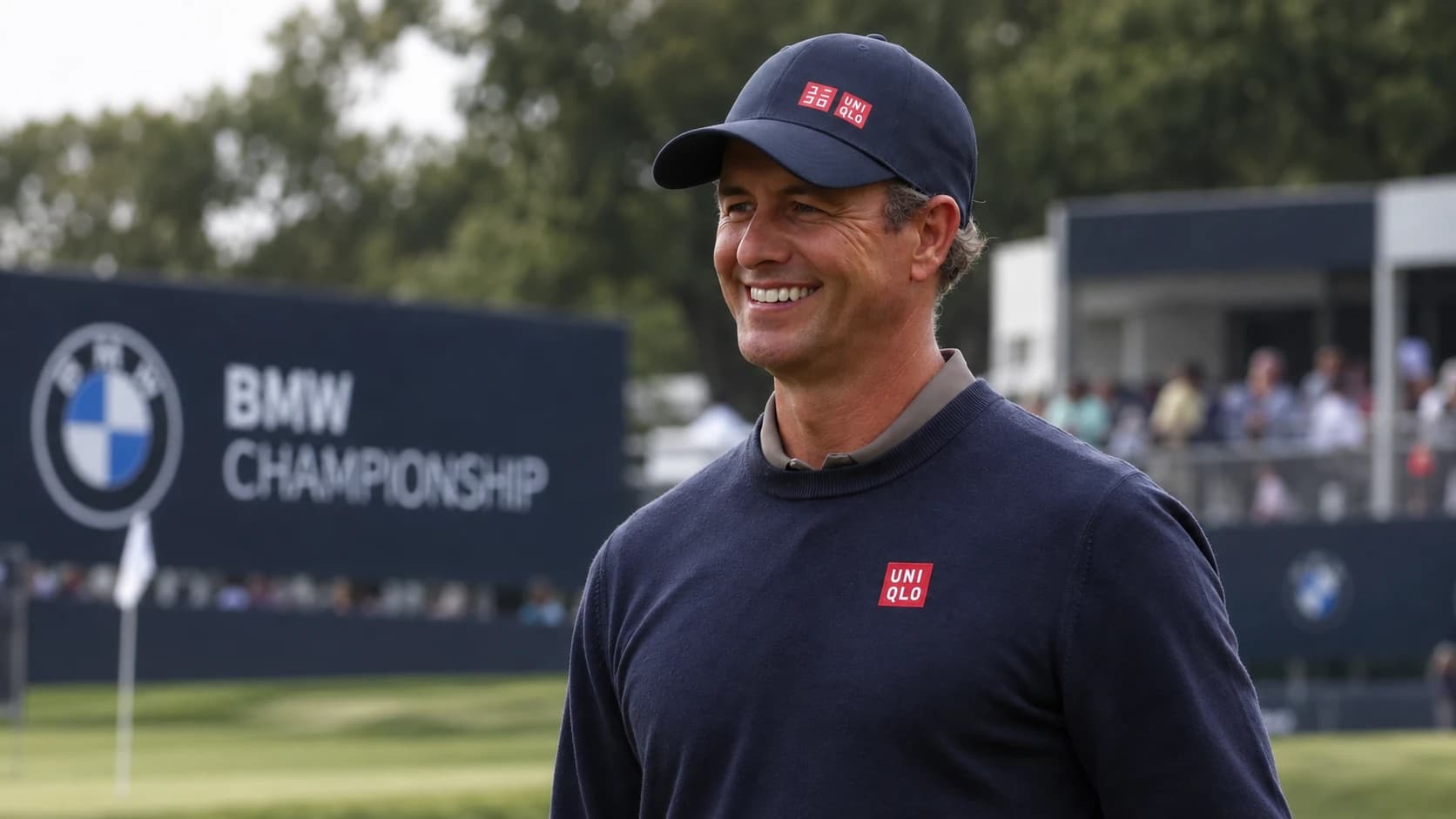 A smiling golfer in a navy sweater and cap on the course, with grandstands in the background